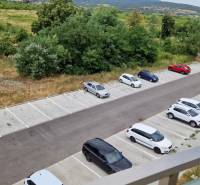 NOVÝ ZARIADENÝ BYT NA Muškáte - Parkovisko pred 2-izbovým bytom na Muškátovej ulici v Pezinku, obklopené zeleňou a kopcami.