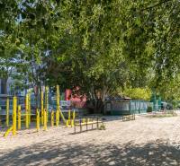 Krásny 3-izbový byt s LODŽINOU a PIVNICOU blízko Draždiak - Park so žltým cvičebným náradím a stromami, pri ulici Budatínska a budovami v pozadí.