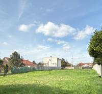 Pozemok 1315 m² pre rodinný dom blízko centra – Sládkovičovo - Pozemok na bývanie v Sládkovičove, tráva, ploty, stromy, budovy v pozadí, oblačná obloha.
