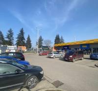 Obchodné priestory na prenájom v nákupnom centre RGB Javorina - Parkovisko pri obchodných priestoroch na ulici Trenčianska s autami a stromami v pozadí.