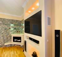 NA PRENÁJOM PRIESTRANNÝ 3-IZBOVÝ BYT V SKVELEJ LOKALITE, UL. SLOBODY - Obývacia stena s televízorom, dekoratívnym kameňom a podlahou s dreveným dekorom v 3-izbovom byte.