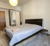 HD REALITY, Vám ponúka na Predaj 3-izbový Apartmán + Garážové státie - Spálňa v 3-izbovom byte s podlahou s dreveným dekorom, posteľou a lampou.