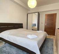 HD REALITY, Vám ponúka na Predaj 3-izbový Apartmán + Garážové státie - Spálňa v 3-izbovom byte s manželskou posteľou, televízorom a podlahou s dreveným dekorom.