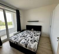 Nový 2 izbový byt SLNEČNICE, Labutia ulica, Bratislava V. Petržalka - Spálňa v 2-izbovom byte s posteľou, skriňou a balkónom s podlahou s dreveným dekorom.