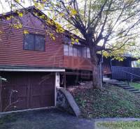 Chata nad kúpeľmi Dudince s vlastným pozemkom - Drevená chata v Dudincoch obklopená stromami a trávnatým svahom.