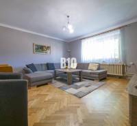 Rodinný dom s krásnym veľkým pozemkom ( stavebným) - Nové Sady - Obývacia izba v rodinnom dome s hnedou podlahou, šedou sedačkou a veľkým oknom.