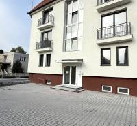 NOVÝ 2 - IZB. BYT (63m2), SENICA - SADOVÁ - Budova na Sadovej ulici v Senici s bielou fasádou a dlaždeným parkoviskom.