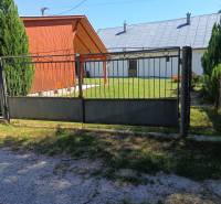 TUŽINA okres Prievidza - CHATA s terasou a garážou  - Brána a drevená chata v Tužine, obklopené trávnikom a ihličnanmi.