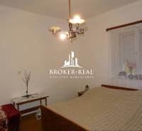 Rodinný dom, chalupa 2 Izbový - Fony - Spálňa v rodinnom dome s posteľou, stolíkom a lustrom, na stene je okno so záclonou.