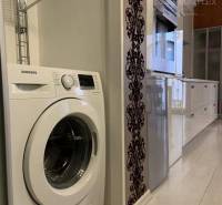 Vám EXKLUZÍVNE ponúka NA PRENÁJOM moderný 2 iz - Kuchyňa v 2-izbovom byte s práčkou, bielym nábytkom a podlahou s dreveným dekorom.