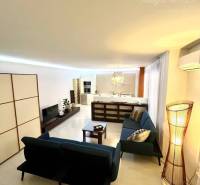 Vám EXKLUZÍVNE ponúka NA PRENÁJOM moderný 2 iz - Obývačka v 2-izbovom byte s podlahou s dreveným dekorom, modrými sedačkami a televízorom.