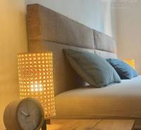 Vám ponúka 1 izbový byt vo VYHĽADÁVANEJ lokali - Čiastočný pohľad na posteľ v 1-izbovom byte s lampou a budíkom.