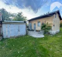 Investičný pozemok s obývateľným domom vo Vrakúni - Rodinný dom vo Vrakúni s plechovou prístavbou obklopený trávnikom a stromami.