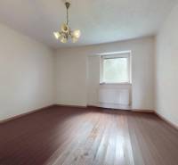 Veľký priestranný pozemok o rozlohe 7445 m2, Jelka  - Izba v rodinnom dome s drevenou podlahou, bielymi stenami a oknom s radiátorom.