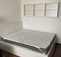 NA PRENÁJOM ÚTULNÝ 1-IZBOVÝ BYT, ČIASTOČNÁ REK., KAROLA ADLERA, BA - Spálňa v 1-izbovom byte s posteľou, bielymi policami a podlahou s dreveným dekorom.