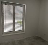 Na predaj dizajnový 4-izbový rodinný dom v Tomášove - Izba v rodinnom dome s veľkým oknom a žalúziami, biele steny, šedá podlaha.