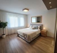 Na predaj priestranný 4-izbový byt 87 m2, Považská Bystrica - Spálňa s posteľou, zrkadlom a nočnými stolíkmi v 4-izbovom byte s podlahou s dreveným dekorom.