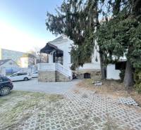 PREŠOV-ŠIRŠIE CENTRUM,POLYFUNKČNÁ BUDOVA,VL.PARKOVISKO,UL.VAJANSKÉHO - Budova na Vajanského v Prešove s prístupovou cestou, autami a smrekmi v popredí.