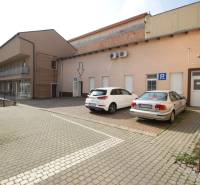 Ponúka na predaj budovu - centrum Senca - NOVÁ SUPER CENA  - Na ulici Mierové nám. pred budovou sú dve autá na parkovisku a smetné koše.