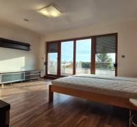 Reprezentatívny 5-izbový rodinný dom pri Piešťanoch - Spálňa v rodinnom dome s podlahou s dreveným dekorom, posteľou a veľkými balkónovými oknami.