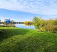 KH-1018, PREDAJ Pozemok pri jazere 887 m², garáž, altánok - Trávnatá plocha a vodná plocha obklopená vegetáciou v Čani. Výstavba domov.