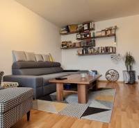 Predaj 2i byt v meste Kremnica, 64m2 - Obývačka v 2-izbovom byte s knihami, šedou sedačkou, kreslom a podlahou s dreveným dekorom.