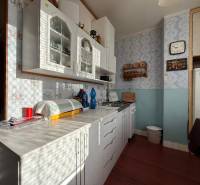 PREDAJ 2 IZBOVÝ BYT HANDLOVÁ  - Kuchyňa 2-izbového bytu so svetlou linkou, poličkou a retro hodinami na stene.