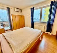 Na predaj – svetlý 2-izbový byt s loggiou a garážou | Bratislava - Spálňa s balkónom, podlahou s dreveným dekorom a modrými závesmi v 2-izbovom byte.