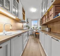 Ponúkame na predaj 3-izb.byt Vígľašská ul., Bratislava - Petržalka. - Úzka kuchyňa s bielymi skrinkami, pultom a podlahou s dreveným dekorom.