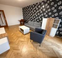 2 izbový byt s balkónom Topoľčany  - Obývačka v 3-izbovom byte s podlahou s dreveným dekorom, vzorovanou tapetou a nábytkom.