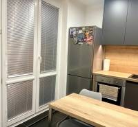 | Predaj 1-izbového bytu – Račianska, Bratislava - Kuchyňa v 1-izbovom byte s drevodekorom, sivý nábytok, veľké okno, chladnička.