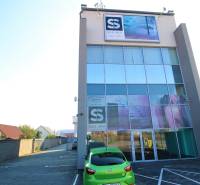 Predaj modernej administratívnej budovy v Bratislave, okres II - Budova so sklenenou fasádou, áutom v popredí a parkoviskom na ulici.