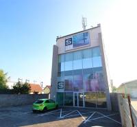 Predaj modernej administratívnej budovy v Bratislave, okres II - Budova s presklenou fasádou a anténou, zaparkované auto, slnečný deň na parkovisku.