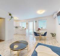 4-izbový byt 92m2 + terasa 14m2 | Bernolákovo - Obývacia izba s kuchyňou, okrúhlym jedálenským stolom a podlahou s dreveným dekorom v 4-izbovom byte.