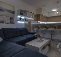 ŠPIČKOVO KOMPLETNE ZARIADENÝ 2-izb. byt bez kompromisov - Obývacia izba s tmavým gaučom, barovými stoličkami a bielymi policami v 2-izbovom byte.