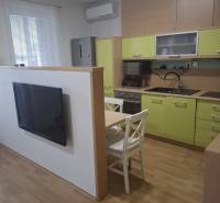 Na prenájom klimatizovaný 2 izbový byt v Moste pri Bratislave - Kuchyňa v 2-izbovom byte so zelenými skrinkami, televízorom a stolom s dvomi stoličkami.