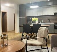 EXKLUZÍVNE-PREDAJ 3-IZBOVÝ BYT POPRAD, širšie centrum 80 m2 s gar.st. - Obývacia izba v 3-izbovom byte s kuchyňou, stolom a podlahou s dreveným dekorom.