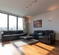 Moderný 2-izbový byt s balkónom, Eurovea - Obývačka v 2-izbovom byte s čiernou sedačkou, podlahou s dreveným dekorom a veľkými oknami.