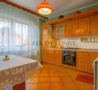 NA PREDAJ 7 IZBOVÝ RODINNÝ DOM, DUNAJSKÁ STREDA - Kuchyňa s drevenými skrinkami, stolom s čipkovým obrusom a podlahou s hnedým dekorom.