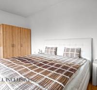 NA PREDAJ APARTMÁNOVÝ DOM VO VEĽKOM MEDERI - Svetlá spálňa s dvojposteľou, šatníkom, nočným stolíkom a podlahou s dreveným dekorom.