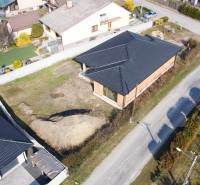 PRIESTRANNÁ 4-IZBOVÁ NOVOSTAVBA 167.38m²- BUNGALOV NA POZEMKU AŽ 702m² - Rodinný dom v Pavliciach pri ceste, so záhradou a susediacimi domami v pozadí.