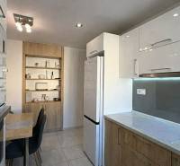 NA PREDAJ vkusne zrekonštruovaný 2 izbový byt, Komárno - Kuchyňa v 2-izbovom byte s bielymi skrinkami, stolom a dreveným dekorom políc.