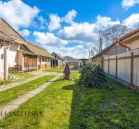 NA PREDAJ 4-IZBOVÝ RODINNÝ DOM NA PRIESTRANNOM POZEMKU - Dvor rodinného domu so záhradou, budovou a stromami na vidieku pod modrou oblohou.