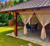 Na predaj 5-izb. rodinný dom na 50-á pozemku, Sap - Záhradná terasa rodinného domu v Sape so stolom, stoličkami a závesmi.