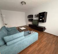 3 izbový byt bez balkóna Topoľčany - Obývacia izba s televízorom, modrou sedačkou a podlahou s dreveným dekorom v 3-izbovom byte.