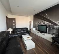 Prenájom 2-izb bytu v Bratislave-Rača - Obývačka 2-izbového bytu s čiernymi pohovkami, podlahou s dreveným dekorom a veľkou tapetou.