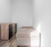 Komfortný 1,5-izbový byt s balkónom v Ružinove na predaj - Úzka miestnosť s posteľou a komodou, podlaha s dreveným dekorom, 1-izbový byt.