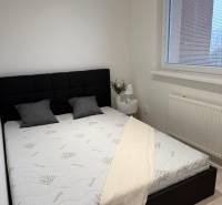Prenájom 2-izbový byt s loggiou, 54m2, Bratislava-Petržalka - Spálňa v 2-izbovom byte s čiernym čelom postele, biela prikrývka a okno s roletami.