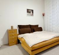 Kompletne zrekonštruovaná garzónka na prenájom, Internátna ulica - Garsónka s posteľou, nočným stolíkom a kobercom, jemné závesy na okne.