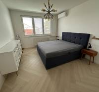 Na predaj moderný 2-izbový byt po kompletnej rekonštrukcii – Piešťany - Spálňa s posteľou, bielou komodou, podlahou s dreveným dekorom a závesným svietidlom.